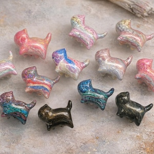 Könnte beinhalten: Eine Sammlung kleiner, katzenförmiger Figuren in verschiedenen Farben. Die Figuren haben ein marmoriertes Aussehen mit Farbtönen von Rosa, Blau, Grün und Schwarz. Jede Katze sitzt mit erhobenem Schwanz.