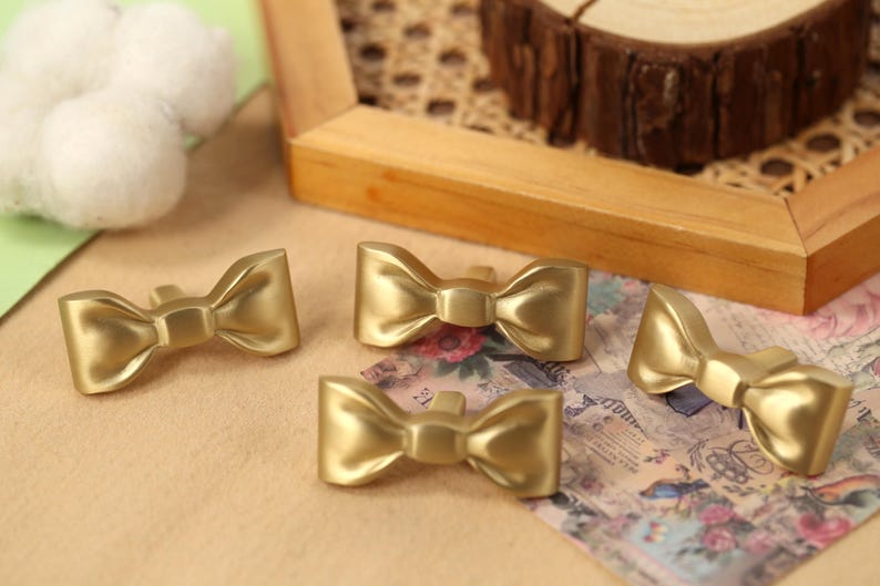 May include: Four gold-coloured bow-shaped drawer pulls. The pulls are made of metal and have a smooth, polished finish.