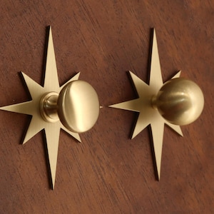 May include: Two brass cabinet knobs, each featuring a circular handle mounted on a flat, eight-pointed star-shaped backplate. The metal has a smooth, brushed finish with a warm gold tone, creating a uniform appearance for both hardware pieces.