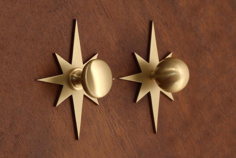 May include: Two brass cabinet knobs, each featuring a circular handle mounted on a flat, eight-pointed star-shaped backplate. The metal has a smooth, brushed finish with a warm gold tone, creating a uniform appearance for both hardware pieces.