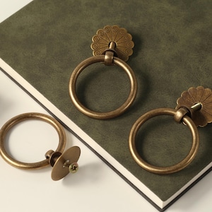 May include: Three antique brass cabinet door pulls. Two of the pulls have a ring shape with a decorative floral design on the top. The third pull has a round ring shape with a flat, round base.