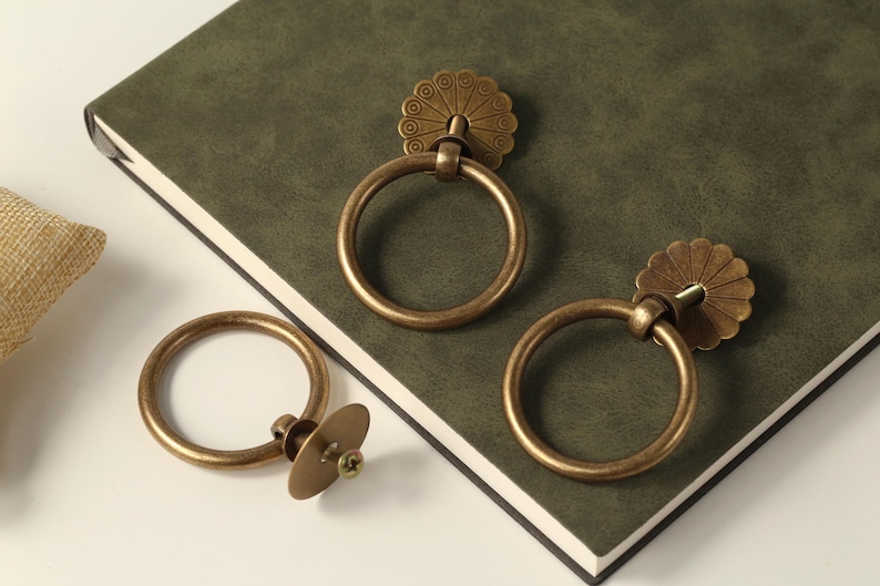 May include: Three antique brass cabinet door pulls. Two of the pulls have a ring shape with a decorative floral design on the top. The third pull has a round ring shape with a flat, round base.