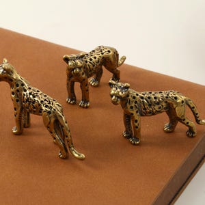 Może przedstawiać: Trzy mosiężne figurki lamparta ze szczegółowym wzorem w cętki. Figurki są ustawione w różnych pozach, z których jedna stoi, jedna kuca, a jedna leży. Figurki znajdują się na brązowej powierzchni.