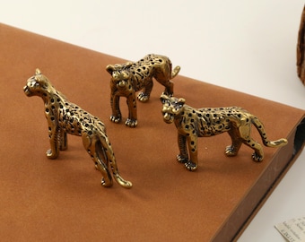 May include: Three brass leopard figurines with a detailed spotted pattern. The figurines are posed in different stances, with one standing, one crouching, and one lying down. The figurines are on a brown surface.