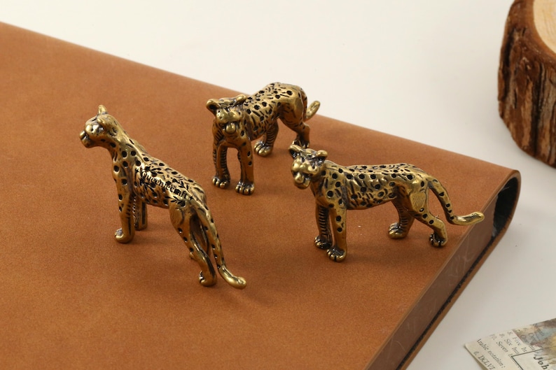 May include: Three brass leopard figurines with a detailed spotted pattern. The figurines are posed in different stances, with one standing, one crouching, and one lying down. The figurines are on a brown surface.