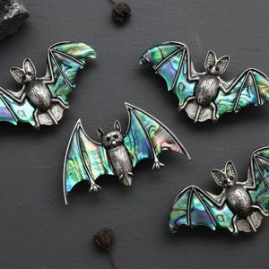 Może przedstawiać: Cztery srebrne broszki w kształcie nietoperzy z iryzującymi skrzydłami w odcieniach zieleni, błękitu i fioletu są prezentowane na ciemnoszarej powierzchni. Nietoperze mają szczegółowe ciała i rozłożone skrzydła.