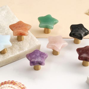 May include: A collection of star-shaped cabinet knobs featuring smooth, polished stone tops in various colors including white, orange, green, black, pink, purple, and maroon, each attached to a small, cylindrical brass-toned metal base.