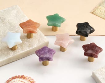 May include: A collection of star-shaped cabinet knobs featuring smooth, polished stone tops in various colours including white, orange, green, black, pink, purple, and maroon, each attached to a small, cylindrical brass-toned metal base.