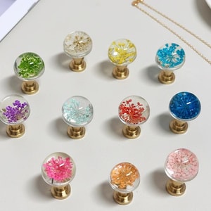Peut inclure: Ensemble de boutons de meubles décoratifs avec des dessus en résine transparente et sphérique. Chaque bouton présente un arrangement floral séché différent dans diverses couleurs, dont le vert, le violet et le rose. Les boutons ont des bases dorées et sont disposés sur une surface blanche.