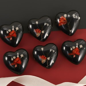 Peut inclure: Six pendentifs en résine noire en forme de cœur avec des fleurs séchées rouges et blanches incrustées. Les pendentifs sont disposés sur une surface rouge et noire avec un ruban blanc.