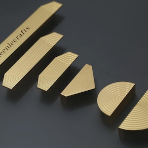 Peut inclure: Ensemble de quincaillerie de placard dorée, comprenant des poignées de barre longues, des formes géométriques et un design semi-circulaire. La quincaillerie présente une texture rainurée et le texte "Queeniecrafts" est gravé.