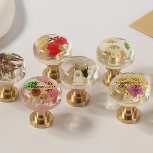 以下が含まれることがあります： 透明な樹脂製のキャビネットノブ7個セット。それぞれ真鍮製の土台と、中にドライフラワーが入った球状のトップが付いています。赤い花、ピンクの花、黄色の花など、さまざまな花の配置があります。「+Queeniecraft+」の文字が一部のノブに見えます。