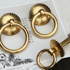 May include: Three gold-toned ring pulls with decorative, scalloped edges. The ring pulls are on a white surface with black text.