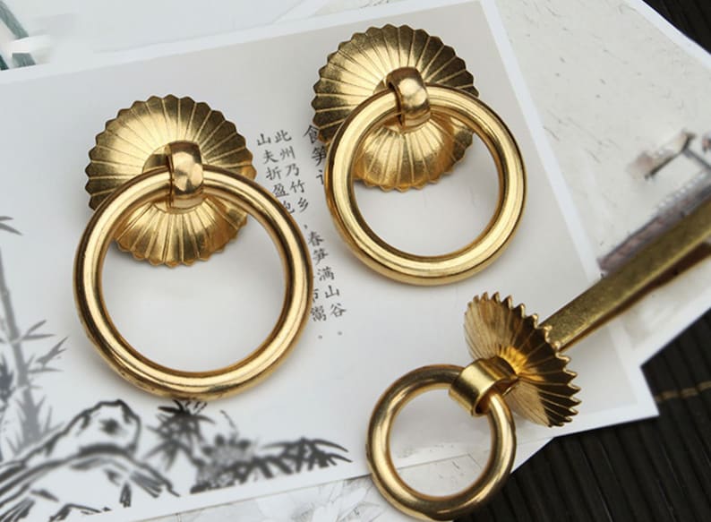 May include: Three gold-toned ring pulls with decorative, scalloped edges. The ring pulls are on a white surface with black text.