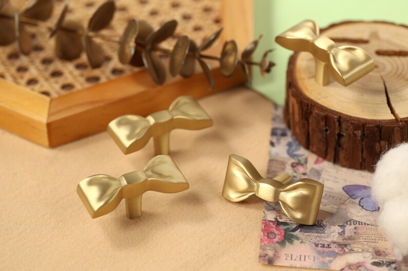 May include: Four gold-toned bow-shaped cabinet knobs with a smooth, shiny finish. The knobs are arranged on a light brown surface.