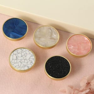 May include: Five round cabinet knobs with gold metal bases and different colored and patterned inserts. The knobs are arranged in a semi-circle on a pink surface. The inserts are blue, white, pink, black, and white with a marble pattern.