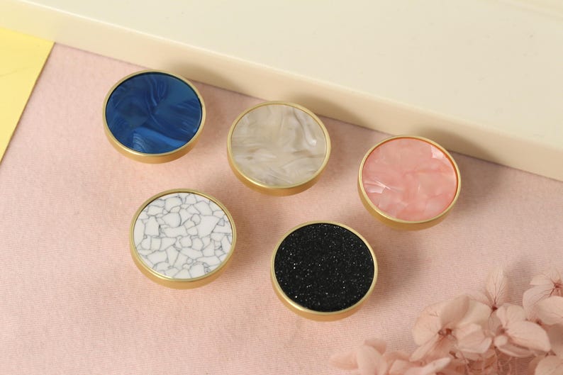 May include: Five round cabinet knobs with gold metal bases and different colored and patterned inserts. The knobs are arranged in a semi-circle on a pink surface. The inserts are blue, white, pink, black, and white with a marble pattern.