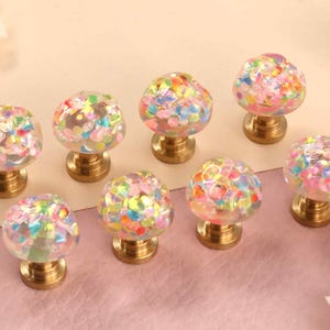 Puede incluir: Un juego de ocho pomos redondos para armarios con tapas transparentes en forma de cúpula incrustadas con copos multicolores e iridiscentes similares a confeti. Cada pomo está unido a una base circular de metal dorado, creando un contraste entre la parte superior transparente y texturizada y los herrajes de metal pulido.