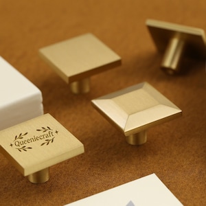 May include: Close-up of several square brass cabinet knobs with a stem. One knob has the text "Queeniecraft +" with a leaf design. The knobs are on a brown surface, near a white object and a decorative plate.