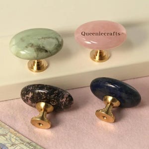Może przedstawiać: Cztery dekoracyjne gałki do szafek z mosiężnymi podstawami i owalnymi kamiennymi blatami. Gałki mają zielone, różowe, czarno-białe i niebieskie wzory kamienne. Różowa gałka ma napis "Queeniecrafts".