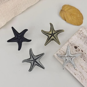 May include: Four starfish-shaped decorative objects in varying metallic finishes: black, gold, silver, and a silver-black blend. The objects are arranged on a neutral surface, with a dried leaf and fabric in the background. These decorative accents are ideal for coastal or nautical themed decor.