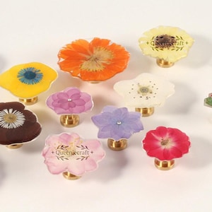Pode incluir: Uma coleção de puxadores decorativos para armários com flores prensadas reais encapsuladas em resina transparente. Os puxadores têm bases de latão e vêm em várias cores, incluindo amarelo, laranja, rosa e roxo. Alguns puxadores têm o texto "+Queeniecraft+".