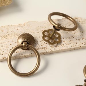 May include: Two antique brass ring pulls with a decorative floral design. The ring pulls are on a white textured surface.
