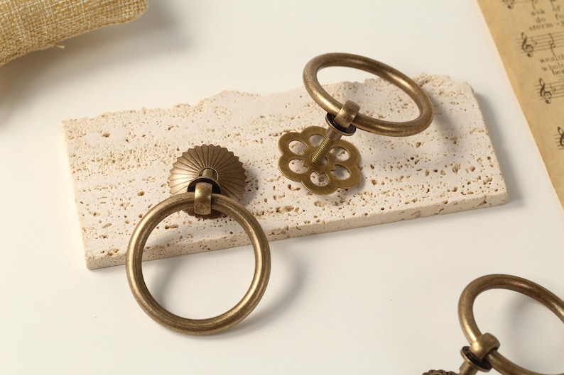 May include: Two antique brass ring pulls with a decorative floral design. The ring pulls are on a white textured surface.