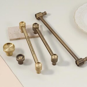 May include: A collection of brass-toned metal cabinet hardware including two round knobs and three long bar pulls. One bar pull features the text Queeniecrafts engraved on its surface. The hardware has a smooth, metallic finish and cylindrical ends.