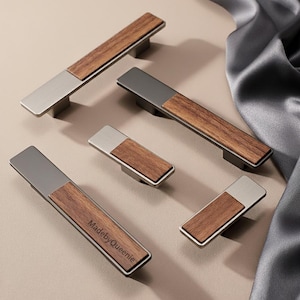 May include: A collection of modern cabinet pulls with a rectangular design. Each pull features a brushed metal end and a wooden center. One pull is labeled "MadebyQueenie". The pulls are arranged on a neutral surface.