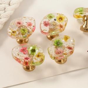 Peut inclure: Des boutons de meubles décoratifs avec des dessus en résine transparente incrustés de fleurs séchées colorées et de paillettes dorées. Les boutons ont des bases de couleur laiton. Un cordon blanc et des roses séchées sont en arrière-plan.