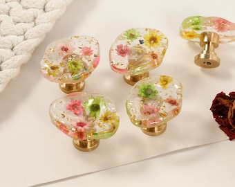 May include: A collection of decorative cabinet knobs with clear resin tops, embedded with colorful dried flowers and gold flakes. The knobs have brass-colored bases. A white rope and dried roses are in the background.
