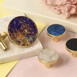 May include: Four decorative cabinet knobs with a brass finish. The knobs feature round, colorful, patterned inlays. The inlays are blue, white, black, and a mix of purple, orange, and gold.