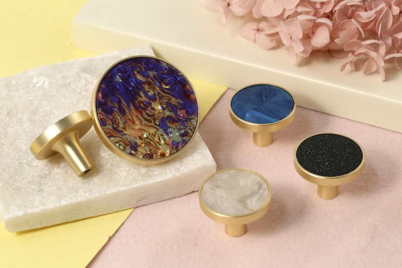 May include: Four decorative cabinet knobs with a brass finish. The knobs feature round, colorful, patterned inlays. The inlays are blue, white, black, and a mix of purple, orange, and gold.