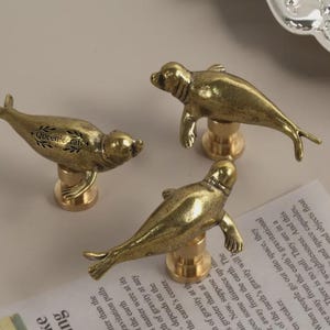 May include: Three antique brass seal-shaped cabinet knobs. Each knob features a detailed seal figure atop a cylindrical base. The knobs are arranged on a light-colored surface, suggesting a decorative or functional hardware item.