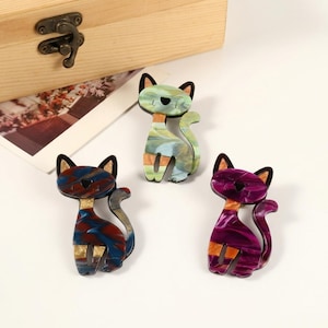 May include: Three colorful cat-shaped brooches. The brooches feature a marbled design in shades of blue, red, green, and purple. Each cat has black ears and a tail. The brooches are made of a shiny material and are set against a white background.