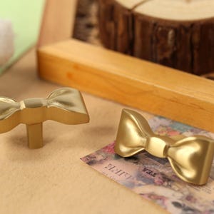 May include: Two gold-coloured bow-shaped cabinet knobs. The knobs are made of metal and have a smooth, shiny finish.