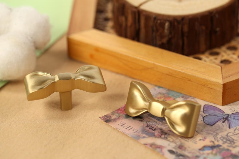 May include: Two gold-coloured bow-shaped cabinet knobs. The knobs are made of metal and have a smooth, shiny finish.