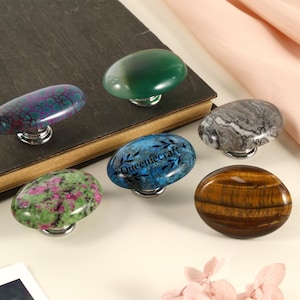 Op de afbeelding: Een verzameling van zes decoratieve kastknoppen met ovale stenen bladen in verschillende kleuren en patronen. De knoppen zijn gemonteerd op zilverkleurige metalen bases. Eén knop heeft de tekst "Queeniecraft".