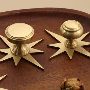 May include: A set of brass cabinet knobs, each featuring a circular top and a tapered base mounted on a flat, multi-pointed star-shaped backplate. The metallic finish has a soft, brushed texture, and the design includes sharp, angular rays.