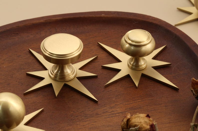 May include: A set of brass cabinet knobs, each featuring a circular top and a tapered base mounted on a flat, multi-pointed star-shaped backplate. The metallic finish has a soft, brushed texture, and the design includes sharp, angular rays.