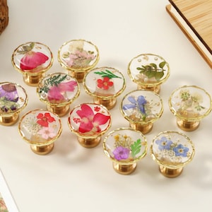 May include: A collection of decorative cabinet knobs with gold-colored bases and clear, round tops. Each knob features a different arrangement of dried flowers encased in resin. The flowers include red, pink, purple, and white varieties.