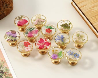 May include: A selection of cabinet knobs with gold-coloured bases and clear, round tops. Each knob features a different arrangement of dried flowers encased in resin. The flowers include red, pink, purple, and white varieties.