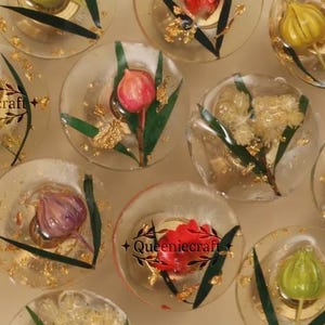 May include: Clear, round resin coasters with embedded flowers and gold flakes. Each coaster features a different flower, including yellow, pink, and red blooms, with green leaves. The coasters are from Queeniecraft.