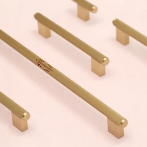 Könnte beinhalten: Goldfarbene Schrankgriffe mit gerilltem Stangendesign und rechteckiger Basis. Die Griffe sind diagonal auf hellem Hintergrund angeordnet. Ein Griff hat den Text "Queenmcraft" in einer dekorativen Schriftart.