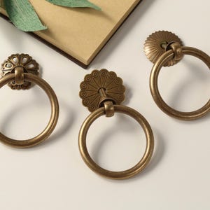 May include: Three antique brass ring pulls with decorative details. The pulls have a circular shape with a ring for pulling. The pulls are perfect for adding a vintage touch to cabinets, drawers, or doors.
