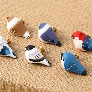 Könnte beinhalten: Sechs handbemalte Vogelfiguren in verschiedenen Farben, darunter Blau, Rot, Braun und Weiß, jeweils mit goldfarbenen Akzenten. Die Vögel sind auf einer strukturierten, beigefarbenen Oberfläche angeordnet. Jedes Vögelchen ist einzigartig.