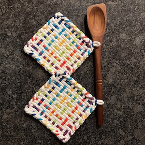 May include: Two square, woven potholders with a white border and a colorful pattern of red, blue, green, yellow, and purple. A wooden spoon rests beside them. The potholders are approximately 7 inches square.