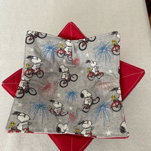 May include: A gray bowl cozy featuring a repeating pattern of Snoopy riding a bicycle with fireworks and stars. The cozy sits atop a red, star-shaped trivet with white stitching. The bowl cozy is designed to keep food warm.