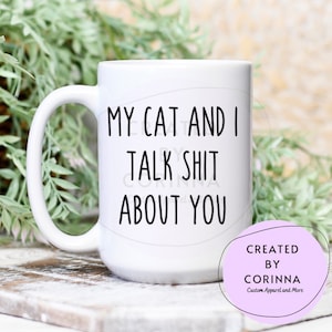 May include: White ceramic coffee mug with black text that reads "My cat and I talk shit about you". The mug is sitting on a wooden surface. The text "Created by Corinna Custom Apparel and More" is visible in a purple circle.